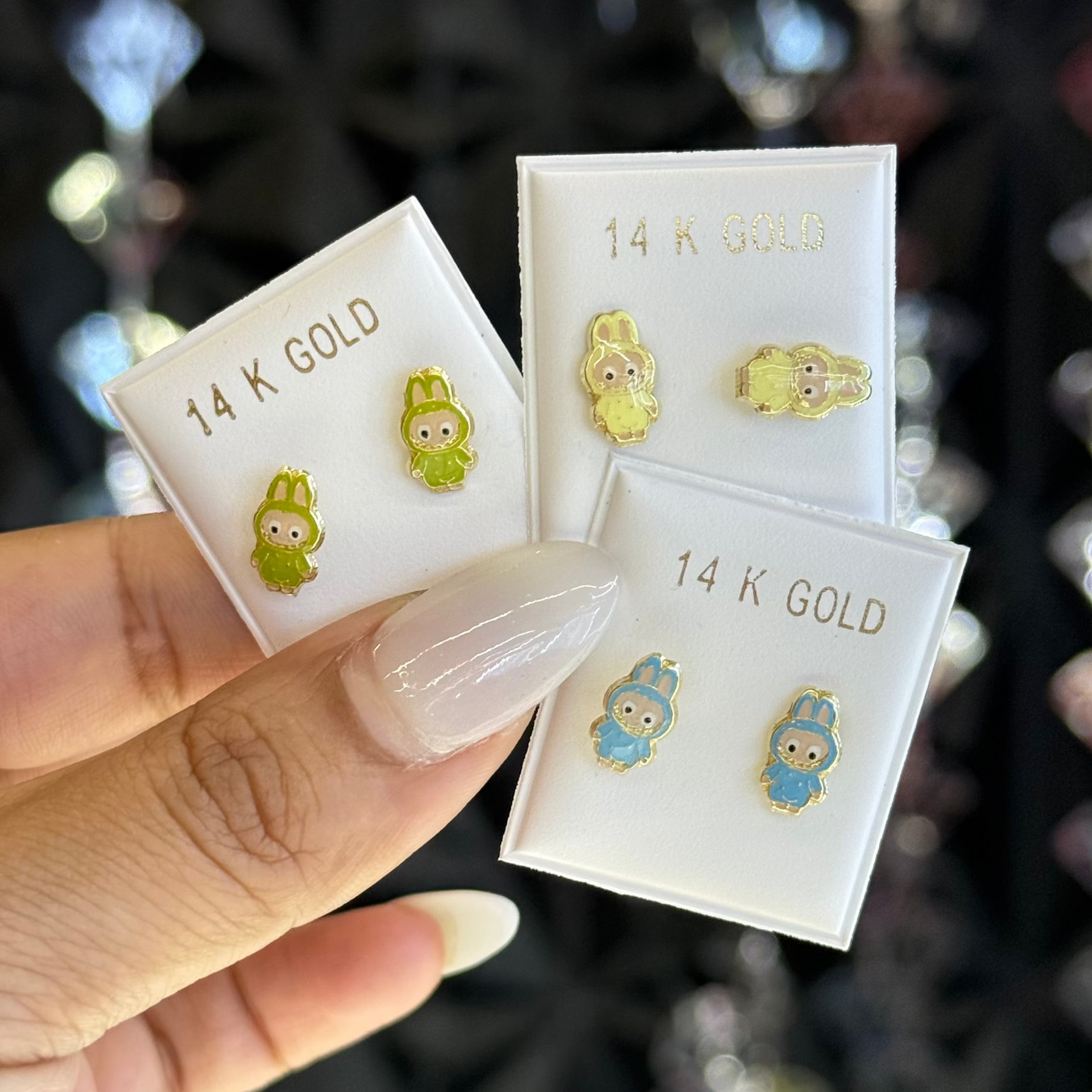 Two pairs of small earrings on '14 K Gold' cards held by a hand against a blurred jewelry store background.