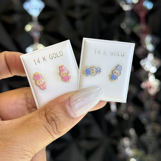 Hand holding two pairs of gold earrings in their packaging with a blurred jewelry store background.